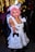 A person with pink hair is dressed in a maid costume, featuring a white dress with black buttons and ruffled edges. The background is festive, with blurred decorative lights and other people visible, some wearing Santa hats.