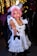 A person with pink hair is dressed in a maid costume, featuring a white dress with black buttons and ruffled edges. The background is festive, with blurred decorative lights and other people visible, some wearing Santa hats.