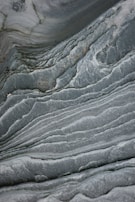 The image features smooth, wavy layers of rock with a textured surface that resembles marble. The swirling patterns create a natural, flowing design marked by variations in gray and green tones.