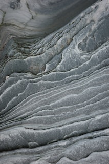 The image features smooth, wavy layers of rock with a textured surface that resembles marble. The swirling patterns create a natural, flowing design marked by variations in gray and green tones.