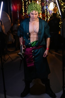 A person dressed in a detailed cosplay costume with green spiky hair and a long dark coat. They have striking makeup accentuating their intense facial expression and hold a sword in one hand. The costume includes a visible scar on their chest and a green sash around their waist. The background is filled with bright lights and blurred figures, suggesting an indoor event.