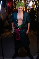 A person dressed in a detailed cosplay costume with green spiky hair and a long dark coat. They have striking makeup accentuating their intense facial expression and hold a sword in one hand. The costume includes a visible scar on their chest and a green sash around their waist. The background is filled with bright lights and blurred figures, suggesting an indoor event.