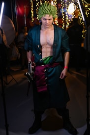 A person dressed in a detailed cosplay costume with green spiky hair and a long dark coat. They have striking makeup accentuating their intense facial expression and hold a sword in one hand. The costume includes a visible scar on their chest and a green sash around their waist. The background is filled with bright lights and blurred figures, suggesting an indoor event.