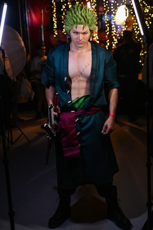 A person dressed in a detailed cosplay costume with green spiky hair and a long dark coat. They have striking makeup accentuating their intense facial expression and hold a sword in one hand. The costume includes a visible scar on their chest and a green sash around their waist. The background is filled with bright lights and blurred figures, suggesting an indoor event.