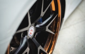 A close-up view of a car wheel showcasing a sleek black alloy rim with a glossy finish and a highlighted orange or bronze lip. The center cap features an emblem with a cross design.