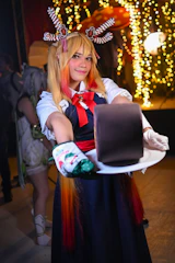 A person in colorful, festive attire presents a chocolate cake on a white plate. The individual has styled blonde and red hair, adorned with decorative accessories resembling candy canes and gingerbread. The background features warm, glowing string lights that create a cozy and celebratory atmosphere.