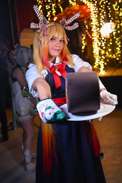 A person in colorful, festive attire presents a chocolate cake on a white plate. The individual has styled blonde and red hair, adorned with decorative accessories resembling candy canes and gingerbread. The background features warm, glowing string lights that create a cozy and celebratory atmosphere.