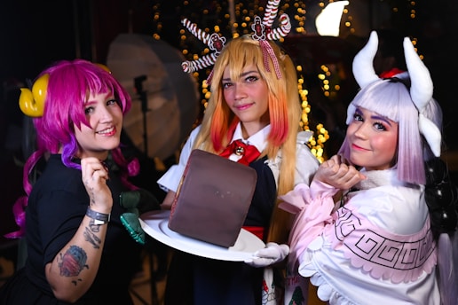 Three individuals are dressed in colorful, elaborate costumes, likely portraying characters from a fantasy or anime setting. They are holding a large rectangular cake on a white plate. The background features blurred lights creating a festive atmosphere.