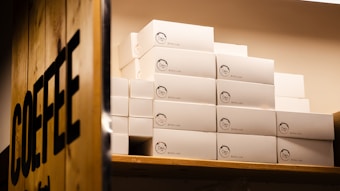 A stack of white boxes with logos are arranged neatly on wooden shelves. To the left, there's a wooden panel with the word 'COFFEE' prominently displayed.