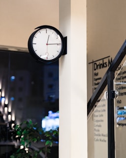 A black-framed wall clock with a white face, black hour and minute hands, and a red second hand is mounted on a light-colored wall. In the background, there is a blurred view of a drinks menu on glass and an interior space with faint lighting visible through a window.