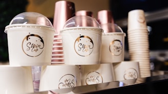 Stacks of disposable coffee cups with translucent lids and logo design. The cups are arranged neatly, featuring a mix of white and pink tones. A blurred background suggests a cafe or food service environment.