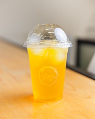 A clear plastic cup filled with a bright yellow drink, likely a juice or soda, is placed on a light wooden surface. The cup has a dome lid and contains ice cubes. There is a logo on the cup that reads 'Stain Cafe'. The background is blurred with neutral tones.
