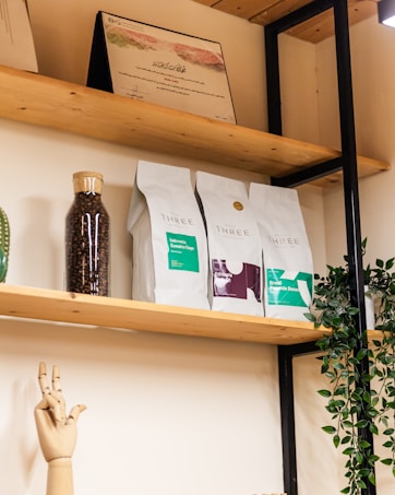 On wooden shelves, there are neatly arranged bags of coffee with labels indicating origins like Sumatra and Ethiopia. A glass jar filled with coffee beans stands next to them. A green plant cascades down from the top shelf, while a wooden mannequin hand is positioned in a gesture. An award or certificate is displayed at the top.