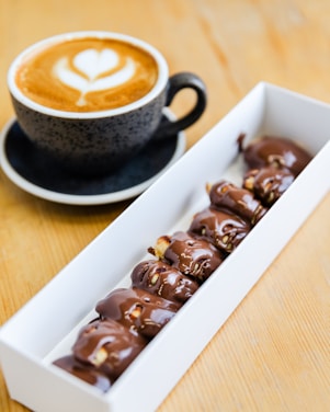 A cup of cappuccino sits on a saucer with intricate latte art on top, accompanied by a rectangular box filled with chocolate-covered pastries. The setting is on a wooden table, giving a warm and inviting feel.