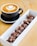 A cup of cappuccino sits on a saucer with intricate latte art on top, accompanied by a rectangular box filled with chocolate-covered pastries. The setting is on a wooden table, giving a warm and inviting feel.
