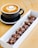 A cup of cappuccino sits on a saucer with intricate latte art on top, accompanied by a rectangular box filled with chocolate-covered pastries. The setting is on a wooden table, giving a warm and inviting feel.