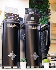 Two black coffee grinders with the brand name 'Victoria Arduino' are prominently displayed. Coffee bags labeled 'Supernatural' and 'Decaf Ethiopia' are placed on top. Small potted succulents are arranged at the base, and there is a leafy plant decor in the background.