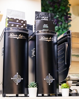 Two black coffee grinders with the brand name 'Victoria Arduino' are prominently displayed. Coffee bags labeled 'Supernatural' and 'Decaf Ethiopia' are placed on top. Small potted succulents are arranged at the base, and there is a leafy plant decor in the background.