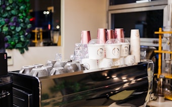 A coffee shop counter displays stacked cups, including white takeaway cups with lids and branded designs, and upside-down ceramic mugs. There's a background of greenery and a window revealing an indoor setting.