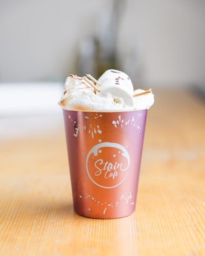 A decorative cup with the logo 'Stain Cafe' holds a dessert topped with whipped cream and marshmallows. The cup has a metallic pink and orange gradient, and is placed on a wooden surface.