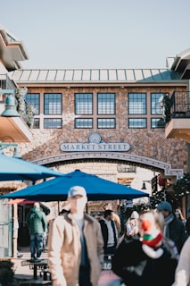 A lively marketplace scene with several people walking around and interacting. The stone facade of a building includes a sign reading 'Market Street' at the entrance. Blue umbrellas are visible in the foreground, with some decorative greenery and festive ornaments enhancing the atmosphere.
