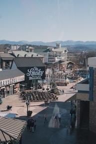 A bustling outdoor shopping area with buildings featuring green roofs and a sign for a distillery or bar. There is a fountain surrounded by seating and people walking around. The backdrop includes mountains under a clear sky.