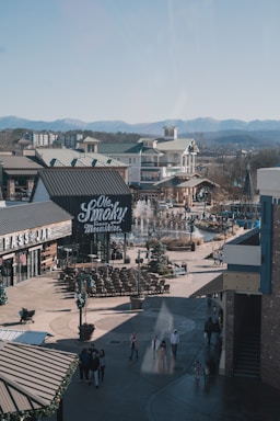 A bustling outdoor shopping area with buildings featuring green roofs and a sign for a distillery or bar. There is a fountain surrounded by seating and people walking around. The backdrop includes mountains under a clear sky.