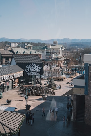 A bustling outdoor shopping area with buildings featuring green roofs and a sign for a distillery or bar. There is a fountain surrounded by seating and people walking around. The backdrop includes mountains under a clear sky.