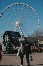 A large Ferris wheel dominates the background, set against a clear blue sky. In the foreground, there is a moonshine store with signage reading 'Ole Smoky'. A couple walks towards the shop, with a decorated Christmas tree visible to the side. The area appears to be an outdoor shopping or entertainment venue.