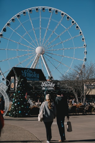 A large Ferris wheel dominates the background, set against a clear blue sky. In the foreground, there is a moonshine store with signage reading 'Ole Smoky'. A couple walks towards the shop, with a decorated Christmas tree visible to the side. The area appears to be an outdoor shopping or entertainment venue.
