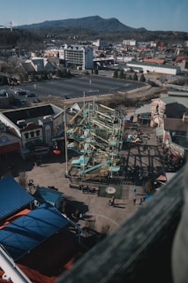 A view from above reveals a theme park with a large, intricate ropes course at the center. Surrounding the course are various buildings, possibly arcade and attraction facilities. Sparse groups of people are visible on the ground, suggesting a relaxed atmosphere. In the background, a range of mountains stretches across the horizon, and several roads and parking lots can be seen, indicating an urban setting nearby.