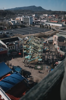 A view from above reveals a theme park with a large, intricate ropes course at the center. Surrounding the course are various buildings, possibly arcade and attraction facilities. Sparse groups of people are visible on the ground, suggesting a relaxed atmosphere. In the background, a range of mountains stretches across the horizon, and several roads and parking lots can be seen, indicating an urban setting nearby.