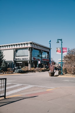 A two-story building with prominent windows and decorative architecture sits on a street corner. Signs on the building indicate a family kitchen theme, and there are winter decorations visible. The area around the building is landscaped with bushes and small trees. Street banners and a pedestrian crosswalk are also present, along with a clear blue sky.