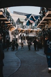 A cozy outdoor shopping area with narrow pathways, flanked by charming storefronts adorned with greenery and decorative elements. People, including families with strollers and children, stroll along the path. The buildings have classic architecture with balconies and awnings, enhancing the quaint atmosphere.