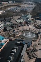 An aerial view of a commercial area featuring a large fountain in the center surrounded by shops and parking lots. The shops have green roofs and there are people walking around the area. The background shows a large parking lot filled with cars and some wooded areas.