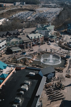An aerial view of a commercial area featuring a large fountain in the center surrounded by shops and parking lots. The shops have green roofs and there are people walking around the area. The background shows a large parking lot filled with cars and some wooded areas.