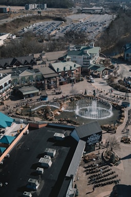 An aerial view of a commercial area featuring a large fountain in the center surrounded by shops and parking lots. The shops have green roofs and there are people walking around the area. The background shows a large parking lot filled with cars and some wooded areas.