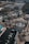 An aerial view of a commercial area featuring a large fountain in the center surrounded by shops and parking lots. The shops have green roofs and there are people walking around the area. The background shows a large parking lot filled with cars and some wooded areas.