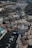 An aerial view of a commercial area featuring a large fountain in the center surrounded by shops and parking lots. The shops have green roofs and there are people walking around the area. The background shows a large parking lot filled with cars and some wooded areas.