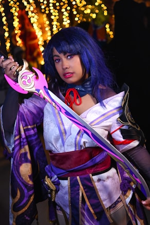 A cosplayer is dressed in a detailed, vibrant costume featuring purple and white fabric with gold accents. The character has blue hair and is holding a decorative sword with a pink and gold hilt. In the background, blurred golden lights create a bokeh effect, adding a festive and enchanting atmosphere.