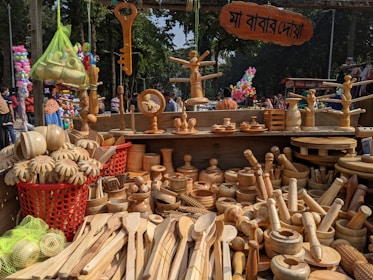A market stall is displaying a variety of wooden kitchen utensils and decorative items. There are wooden spoons, bowls, rolling pins, and other handcrafted items meticulously laid out. Above the stall, a sign with text in a local script is visible. In the background, trees and people are seen milling about, indicating that the market is held outdoors. The vibrant atmosphere is enhanced by the presence of colorful balloons and other stalls in the background.