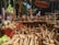 A market stall is displaying a variety of wooden kitchen utensils and decorative items. There are wooden spoons, bowls, rolling pins, and other handcrafted items meticulously laid out. Above the stall, a sign with text in a local script is visible. In the background, trees and people are seen milling about, indicating that the market is held outdoors. The vibrant atmosphere is enhanced by the presence of colorful balloons and other stalls in the background.