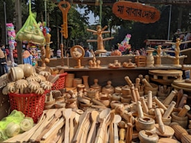 A market stall is displaying a variety of wooden kitchen utensils and decorative items. There are wooden spoons, bowls, rolling pins, and other handcrafted items meticulously laid out. Above the stall, a sign with text in a local script is visible. In the background, trees and people are seen milling about, indicating that the market is held outdoors. The vibrant atmosphere is enhanced by the presence of colorful balloons and other stalls in the background.