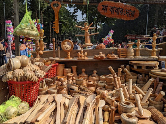 A market stall is displaying a variety of wooden kitchen utensils and decorative items. There are wooden spoons, bowls, rolling pins, and other handcrafted items meticulously laid out. Above the stall, a sign with text in a local script is visible. In the background, trees and people are seen milling about, indicating that the market is held outdoors. The vibrant atmosphere is enhanced by the presence of colorful balloons and other stalls in the background.