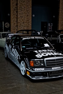 A black sports car with racing decals and a pronounced front grille is parked indoors. The vehicle features numerous logos including AMG, Bridgestone, and Shell, and has white wheels and sleek aerodynamic design. Behind the car, there is a brick wall and a partially obscured classic car.