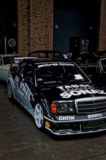 A black sports car with racing decals and a pronounced front grille is parked indoors. The vehicle features numerous logos including AMG, Bridgestone, and Shell, and has white wheels and sleek aerodynamic design. Behind the car, there is a brick wall and a partially obscured classic car.