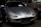 A sleek, silver sports car is parked indoors among other vintage vehicles. The car has a low profile, smooth curves, and oval headlights, giving it a classic and stylish appearance. The badge on the front hood and the number plate indicate it is part of an automobile collection.
