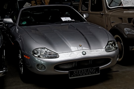 A sleek, silver sports car is parked indoors among other vintage vehicles. The car has a low profile, smooth curves, and oval headlights, giving it a classic and stylish appearance. The badge on the front hood and the number plate indicate it is part of an automobile collection.