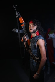 A man with curly hair, wearing a red headband and camouflage sleeveless shirt, is holding an assault rifle. He has an ammunition belt slung across his torso and is looking directly at the camera with a serious expression. Another person in a colorful outfit is slightly visible in the background.