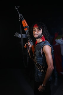 A man with curly hair, wearing a red headband and camouflage sleeveless shirt, is holding an assault rifle. He has an ammunition belt slung across his torso and is looking directly at the camera with a serious expression. Another person in a colorful outfit is slightly visible in the background.